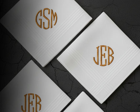 Three white cotton men's handkerchiefs with gold embroidered initials on a dark textured background