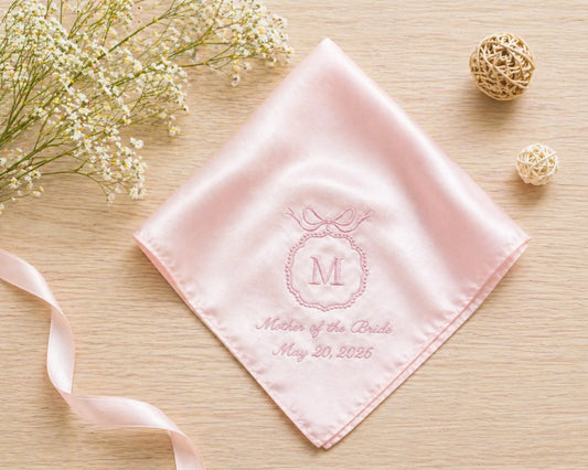 Pink handkerchief with monogram and text on a wooden surface with flowers and ribbons.