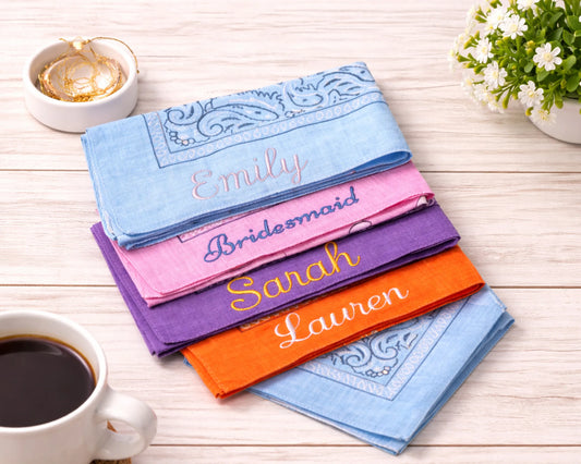 Set of personalized handkerchiefs with names embroidered on them, placed on a wooden surface with a cup of coffee and a plant.