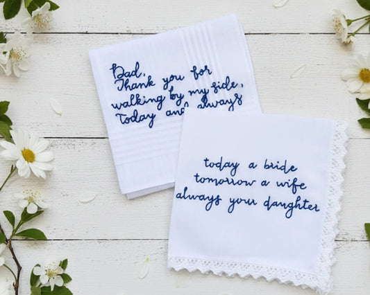Two white handkerchiefs with embroidered text on a wooden surface with flowers.