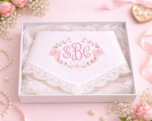 White handkerchief with 'SBe' monogram in a decorative box on a pink background with flowers and jewelry.