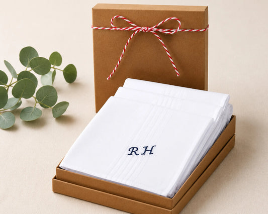 White folded fabric with initials 'RH' in a brown box with a red and white ribbon, on a beige surface with green leaves.