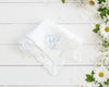 Single white cotton women's hanky with a blue embroidered letter M and decorative lace border next to daisies