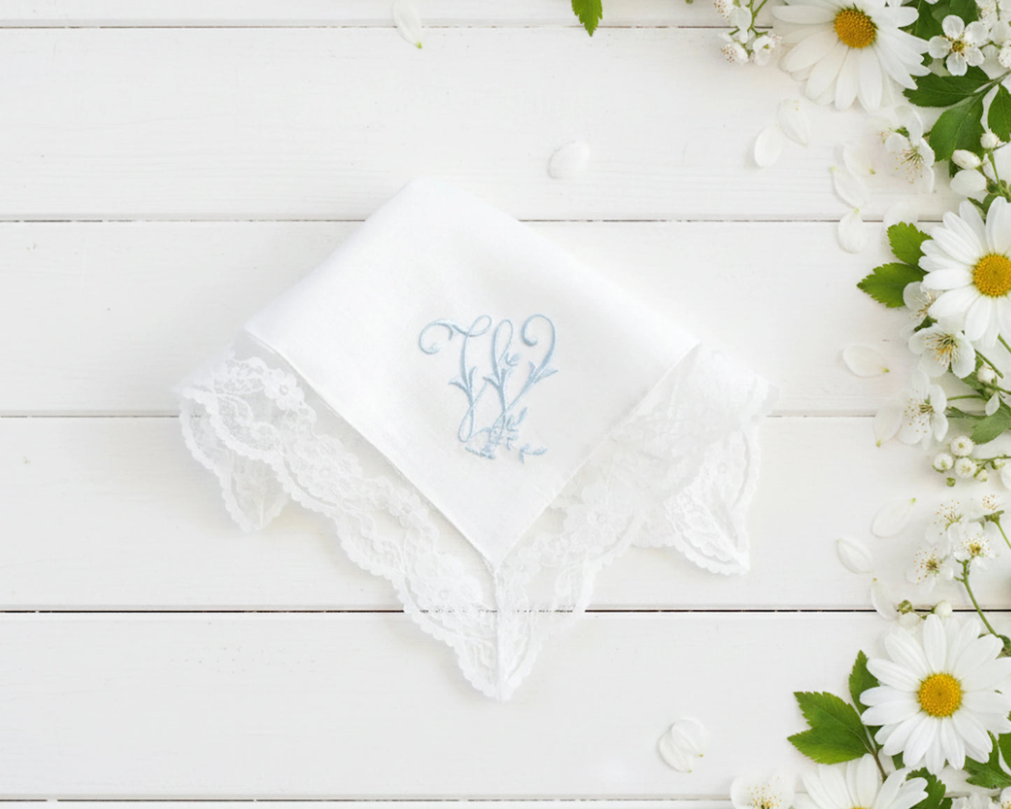 Single white cotton women's hanky with a blue embroidered letter M and decorative lace border next to daisies