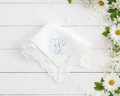 Single white cotton women's hanky with a blue embroidered letter M and decorative lace border next to daisies