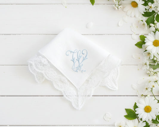 Single white cotton women's hanky with a blue embroidered letter M and decorative lace border next to daisies