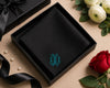 Black box with a monogram on a beige surface with flowers and ribbons.