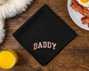 Black napkin with 'DADDY' embroidery on a wooden table with breakfast items.