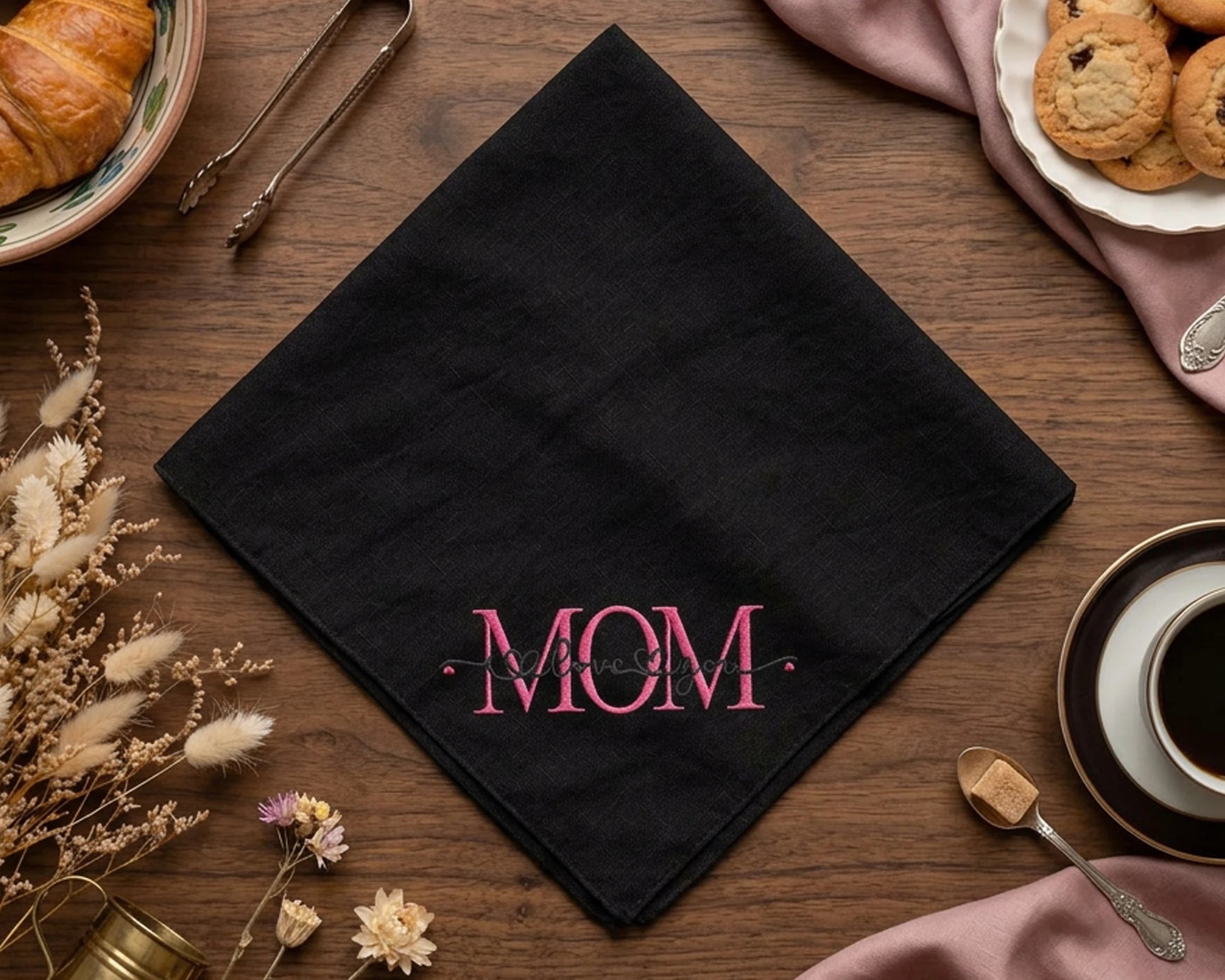 Black napkin with 'MOM' embroidery on a wooden table with coffee and cookies.