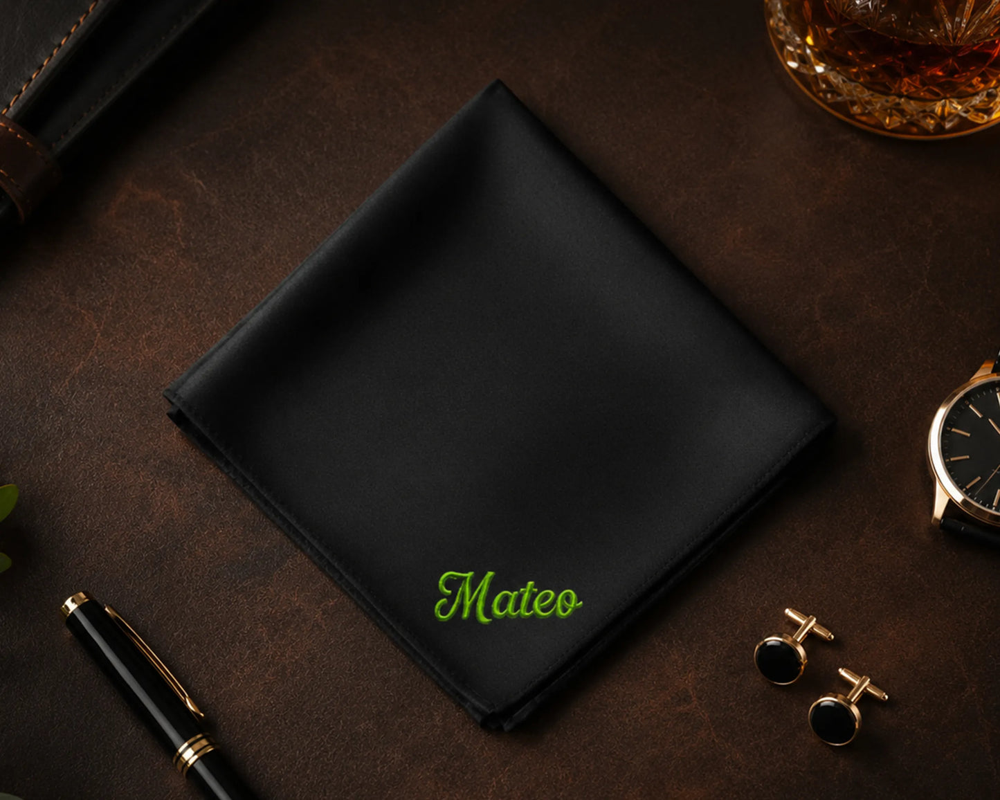 Black pocket square with 'Mateo' embroidery on a dark surface with a watch, pen, and cufflinks.