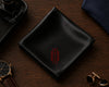 Black pocket square with a red monogram on a wooden surface with accessories.