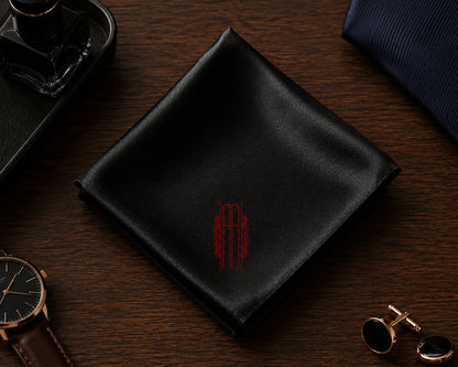 Black pocket square with a red monogram on a wooden surface with accessories.