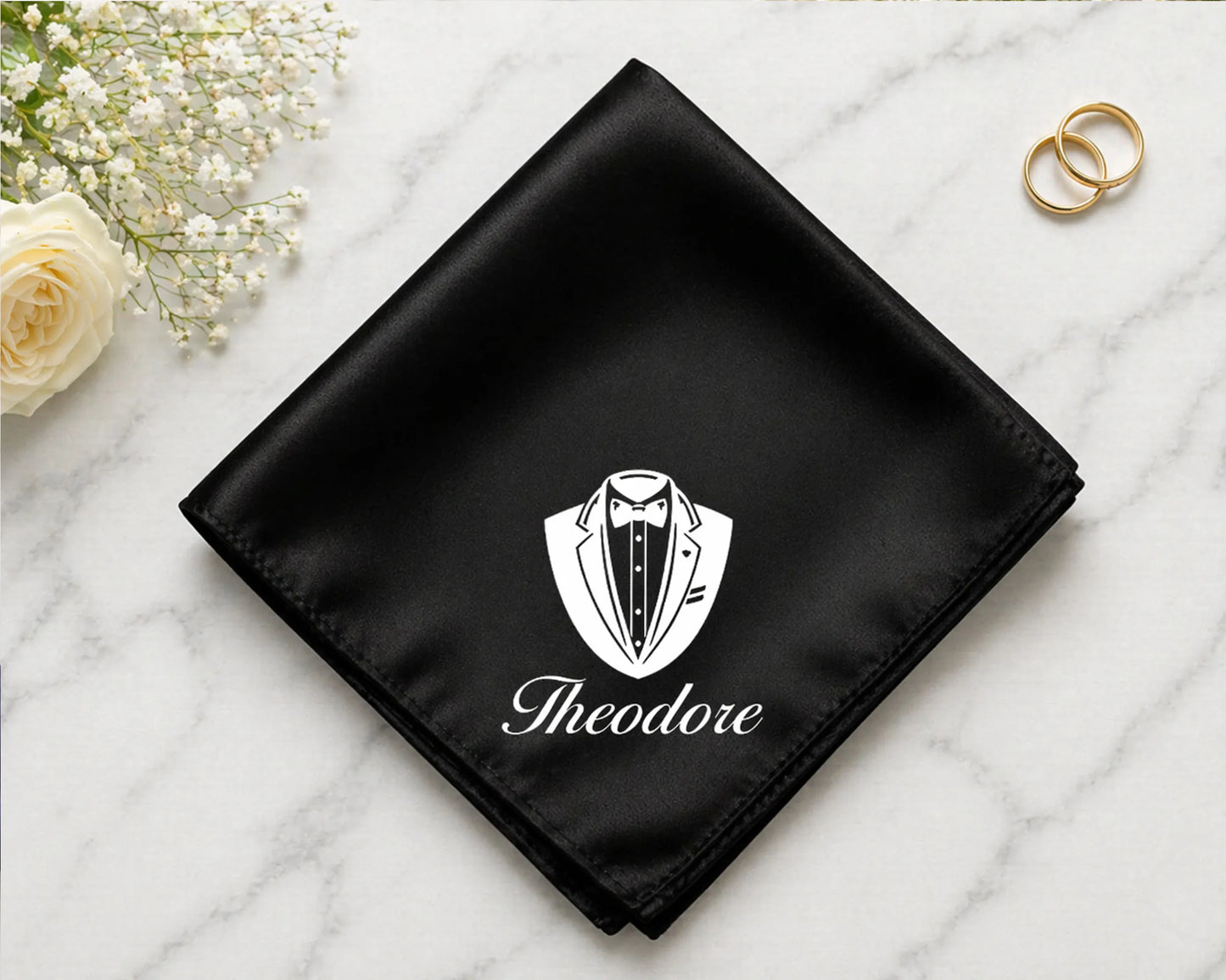Black pocket square with a white emblem and 'Theodore' text on a marble surface with flowers and rings.