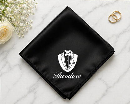 Black pocket square with a white emblem and 'Theodore' text on a marble surface with flowers and rings.