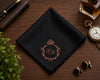 Black pocket square with monogram, pen, wallet, perfume bottle, and pocket watch on a wooden surface.