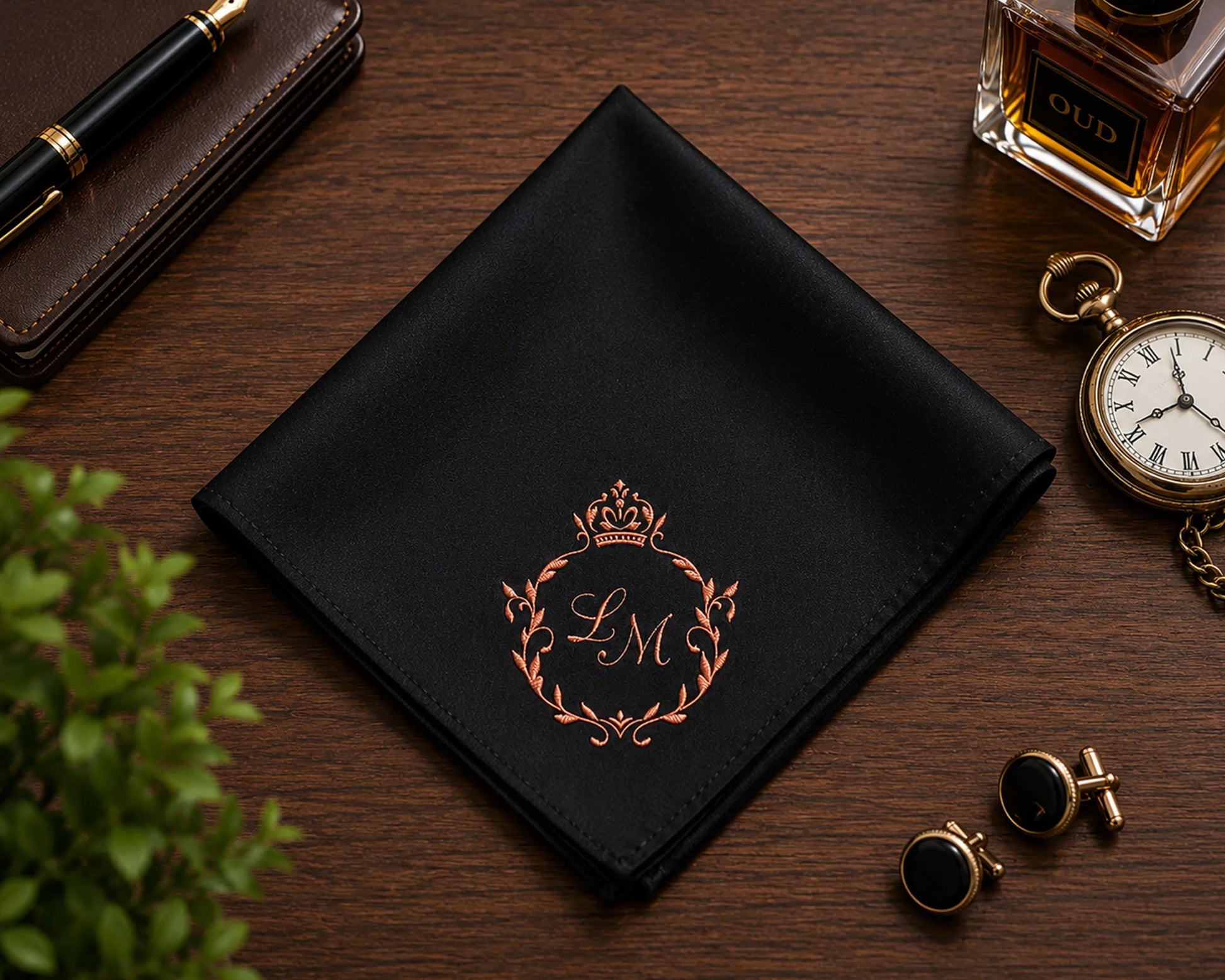 Black pocket square with monogram, pen, wallet, perfume bottle, and pocket watch on a wooden surface.