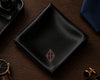 Black satin pocket square with a logo on a wooden surface, surrounded by accessories.