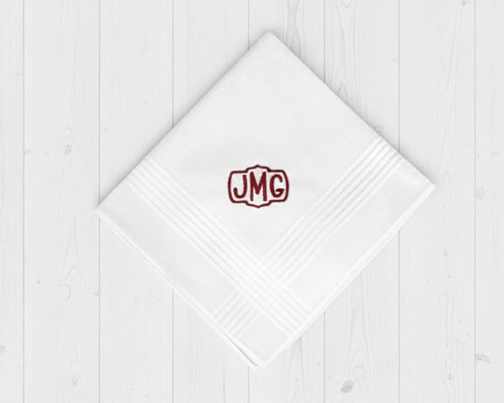 Single white cotton hanky for men with a burgundy embroidered crest monogram JMG on white wood