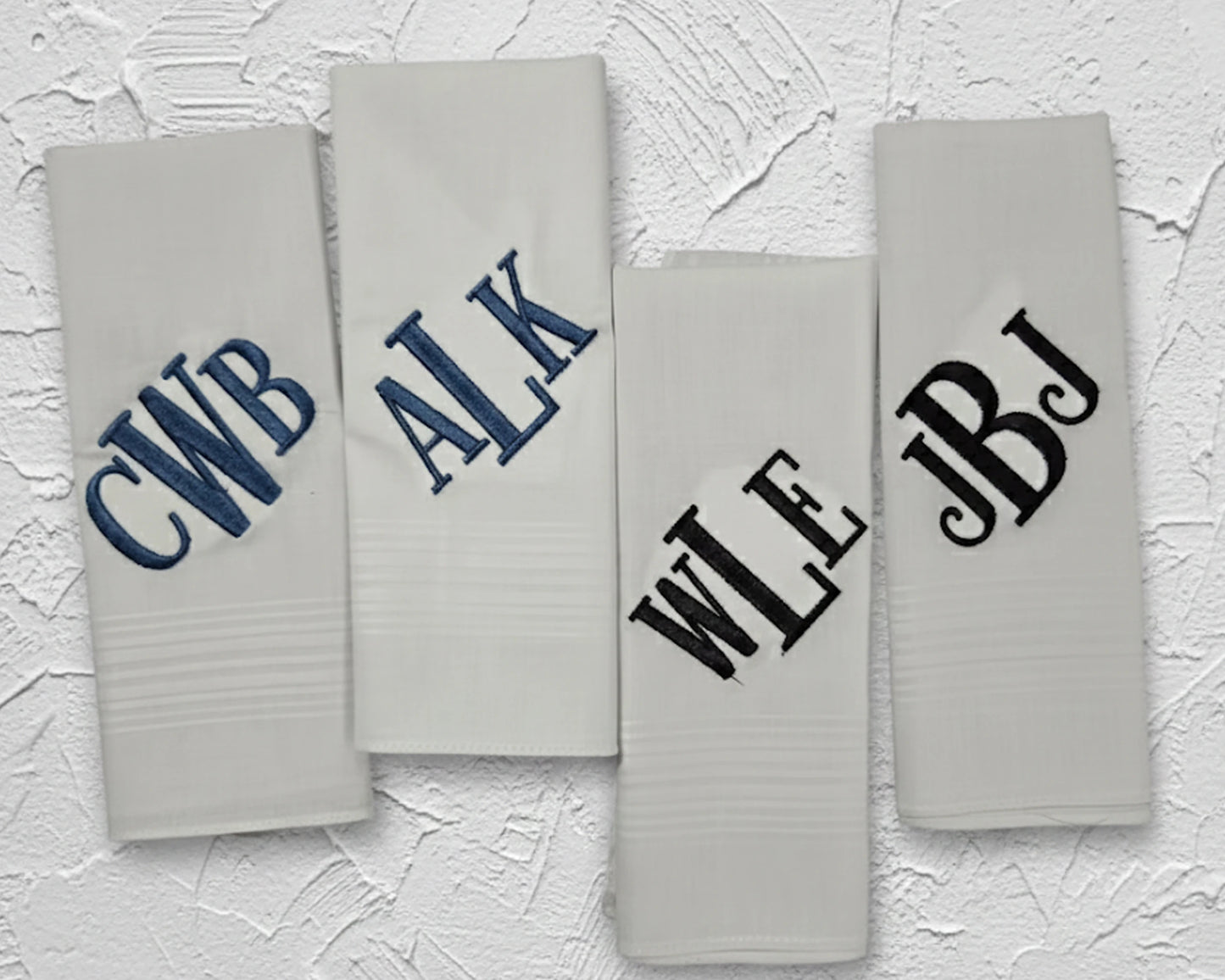 Four folded white cotton handkerchiefs showing diverse navy blue and black monogrammed initial embroidery