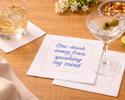 Cocktail glass with a garnish on a coaster with text, next to a glass of iced drink and a bowl of snacks on a wooden table.