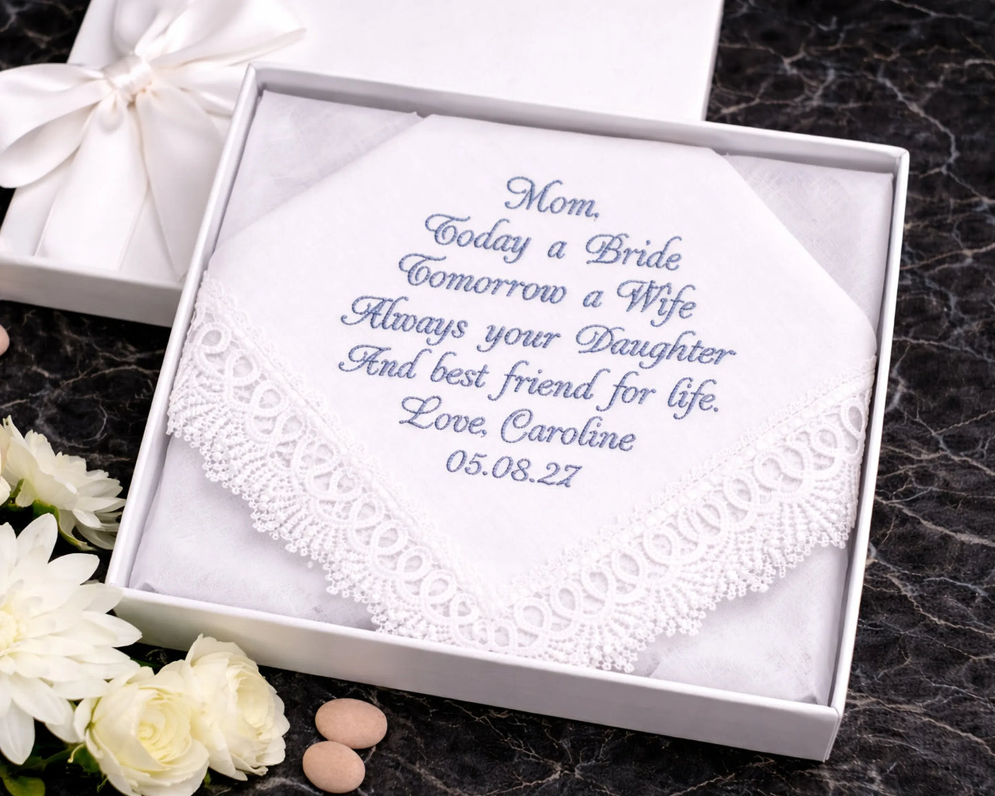 Embroidered handkerchief in a white box with personalized text on a dark surface with flowers.