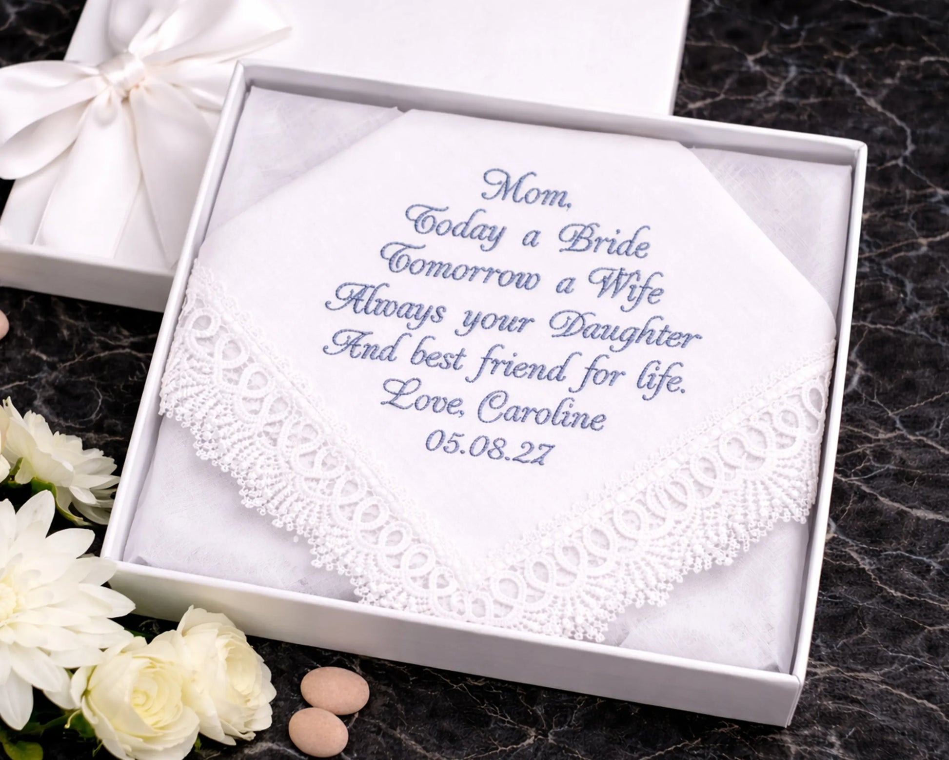 Embroidered handkerchief in a white box with personalized text on a dark surface with flowers.
