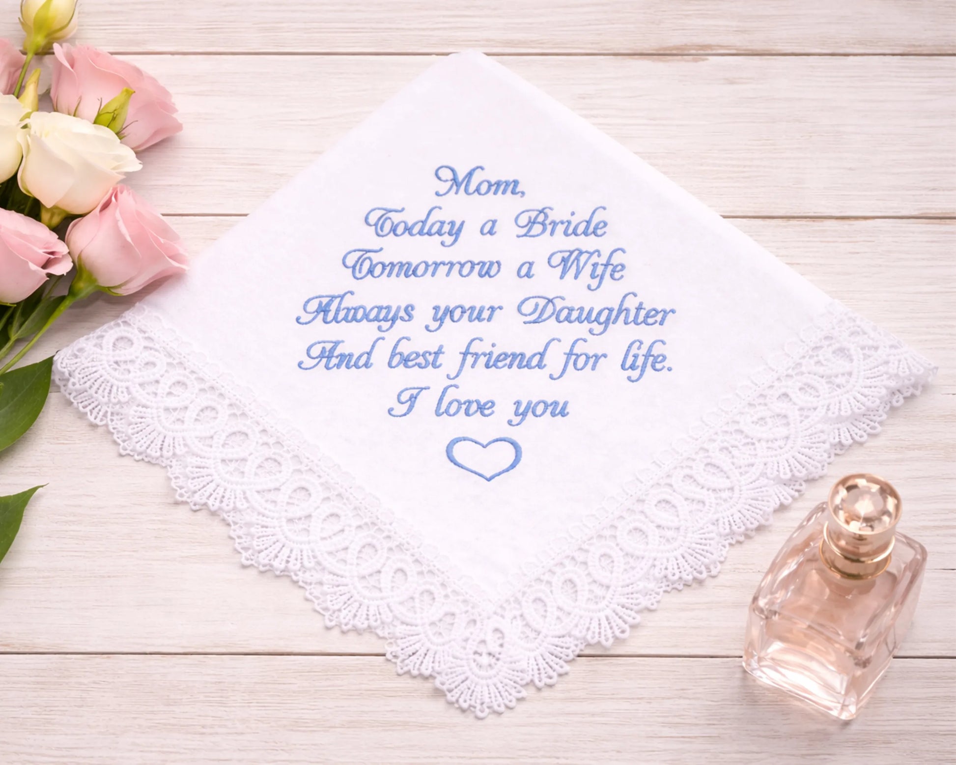 Embroidered handkerchief with a message on a wooden surface with flowers and a perfume bottle.