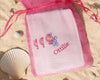 Embroidered hanky with cartoon characters and name 'Ottilie' in a pink drawstring bag on sand with shells.