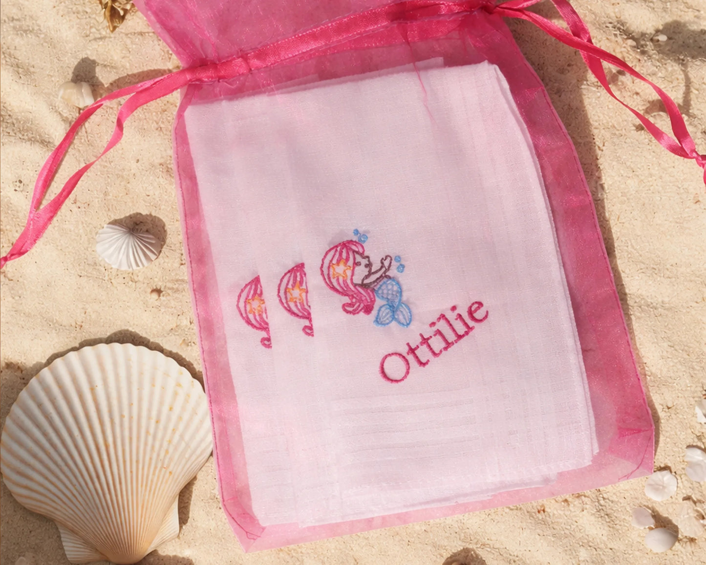 Embroidered hanky with cartoon characters and name 'Ottilie' in a pink drawstring bag on sand with shells.