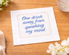 Embroidered napkin with text 'One drink away from speaking my mind' on a wooden surface with flowers.