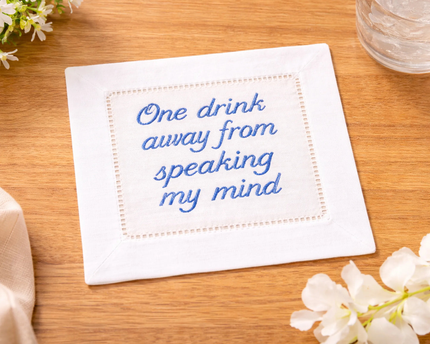 Embroidered napkin with text 'One drink away from speaking my mind' on a wooden surface with flowers.