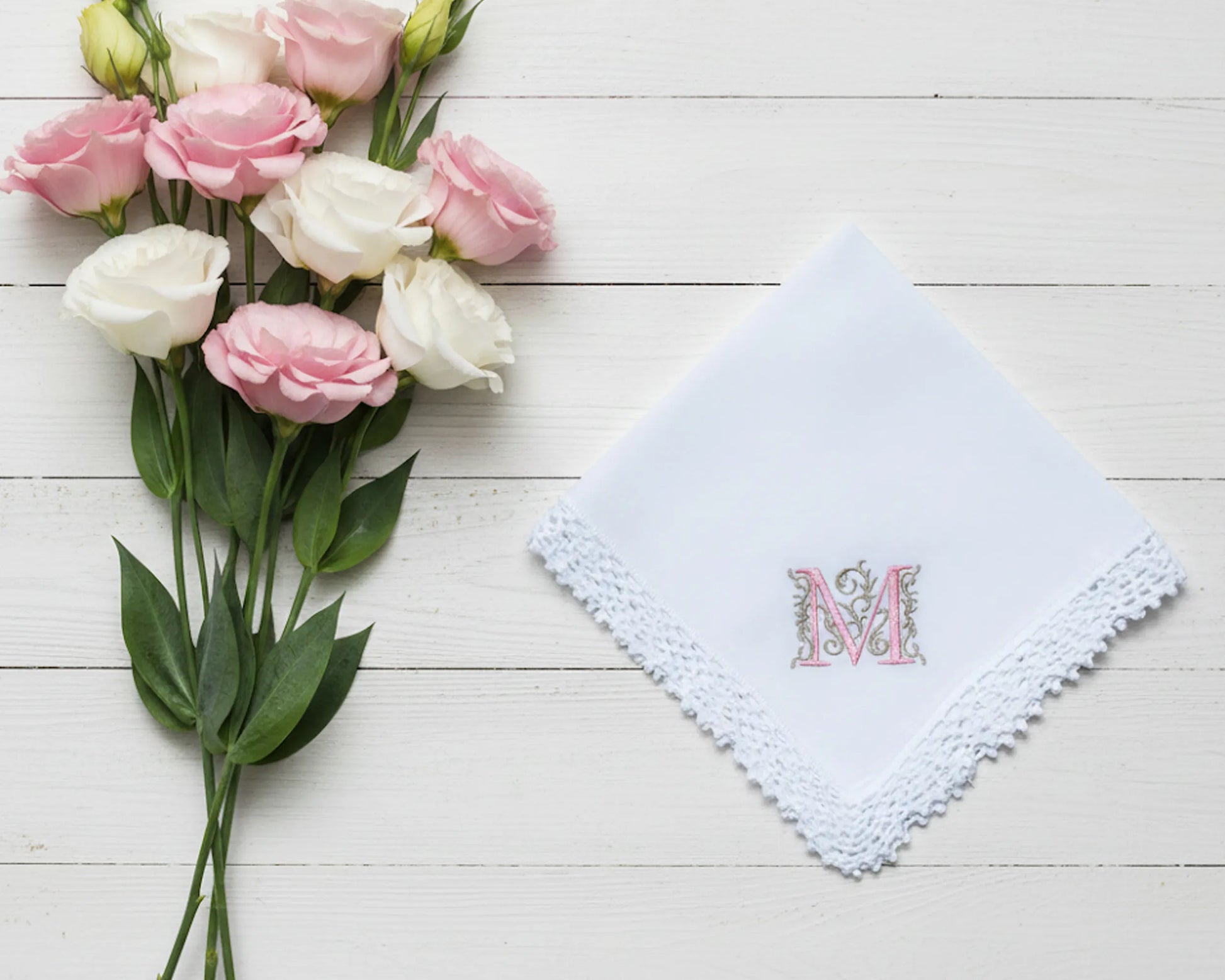 A white cotton handkerchief with a floral M monogram and white lace trim placed next to pink and white roses