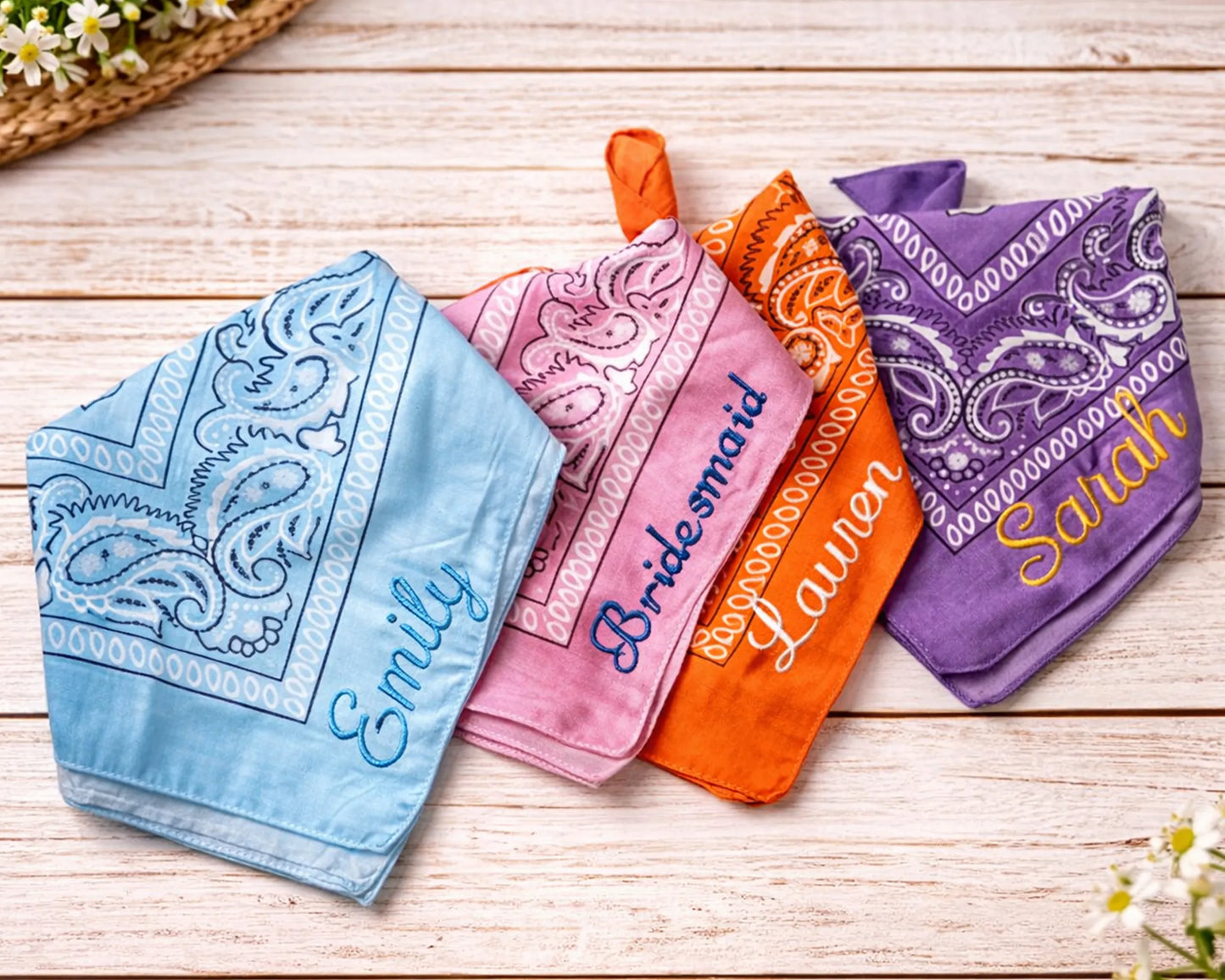 Four bandanas in blue, pink, orange, and purple with embroidered names on a wooden surface.