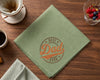 Green hanky with 'Best Dad Ever' embroidery on a wooden table with coffee and berries.