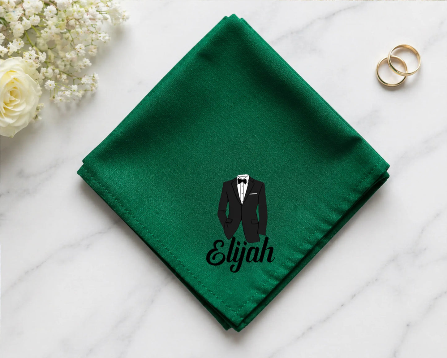 Green hanky with a tuxedo and 'Elijah' text on a white surface with flowers and rings.