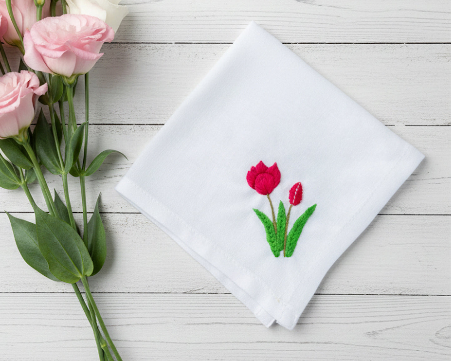 White cotton handkerchief decorated with vibrant red tulip needlework shown next to fresh pink roses