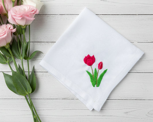 White cotton handkerchief decorated with vibrant red tulip needlework shown next to fresh pink roses