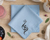Light blue napkin with a black symbol on a beige surface with a glass of pink drink and bowl of fruit.