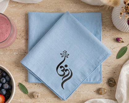 Light blue napkin with a black symbol on a beige surface with a glass of pink drink and bowl of fruit.