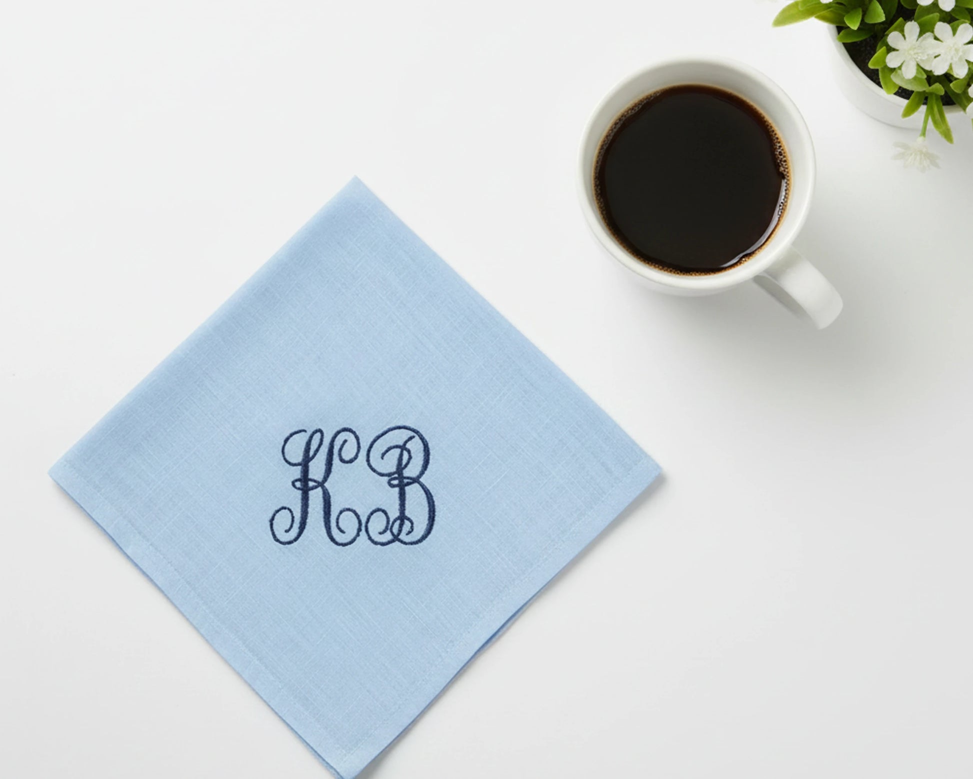 Light blue napkin with monogram 'KB' next to a cup of coffee on a white surface.