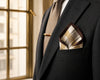 Man wearing a black suit with a patterned pocket square and brown tie, standing in front of a window.