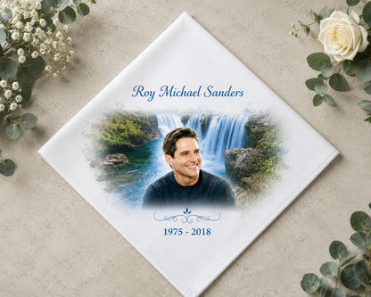 Memorial hank with a photo of a man and a waterfall, surrounded by flowers on a beige surface. 