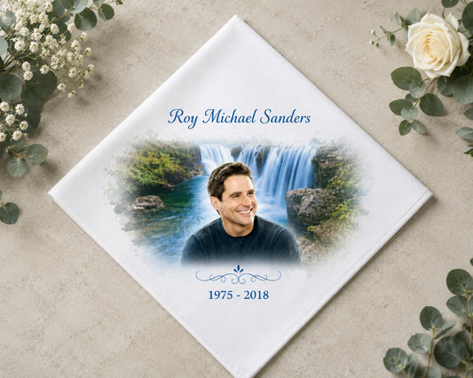 Memorial hank with a photo of a man and a waterfall, surrounded by flowers on a beige surface. 