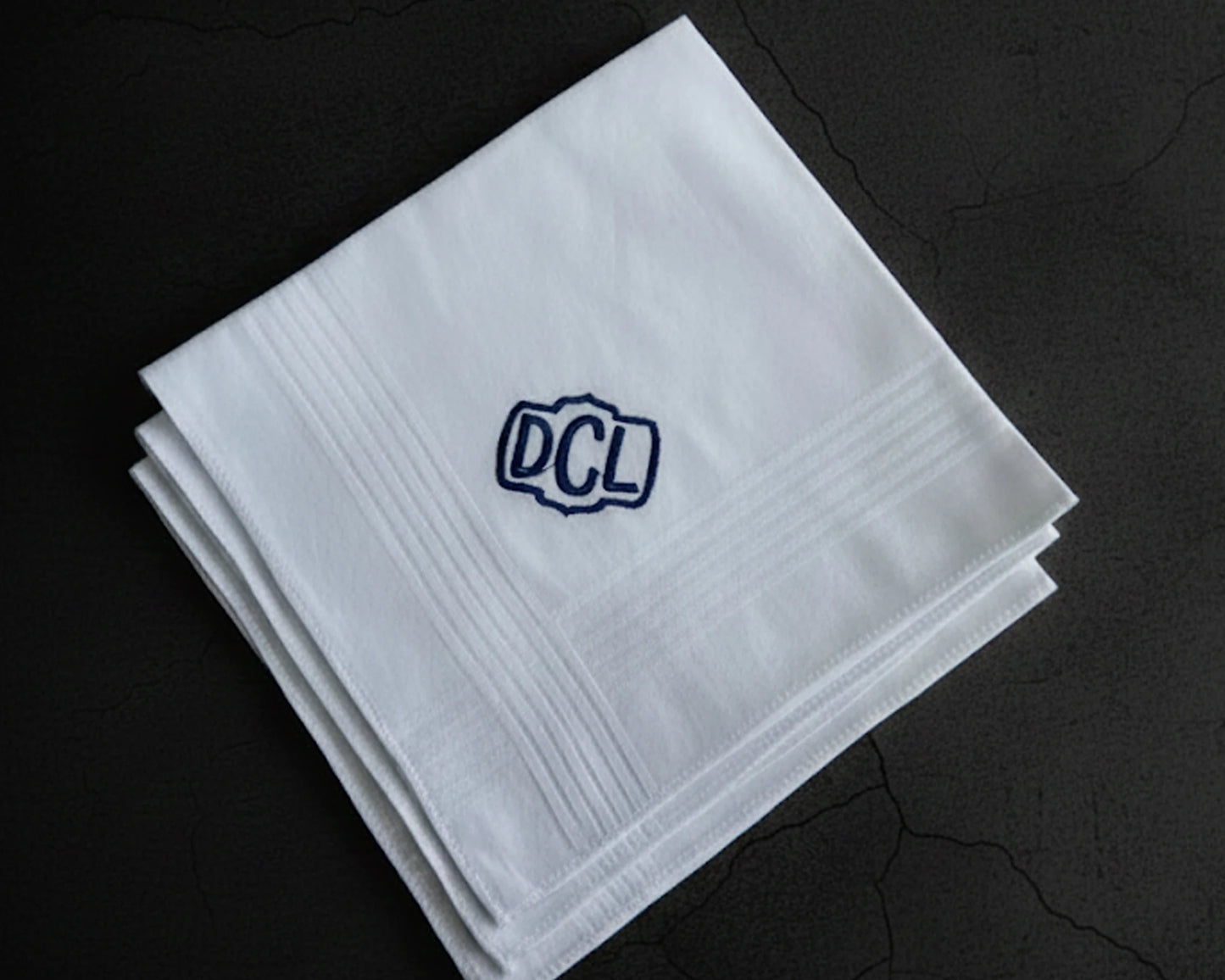 Three white cotton men's handkerchiefs featuring a navy blue embroidered shield monogram DCL on a dark background