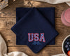 Navy blue napkin with 'USA' embroidery on a wooden table with a glass of pink drink and bowl of berries.