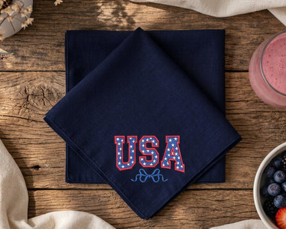 Navy blue napkin with 'USA' embroidery on a wooden table with a glass of pink drink and bowl of berries.