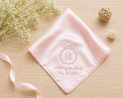 Pink handkerchief with monogram and text on a wooden surface with flowers and ribbons.