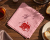 Pink hanky with a red bull logo on a wooden table with tea and bread.