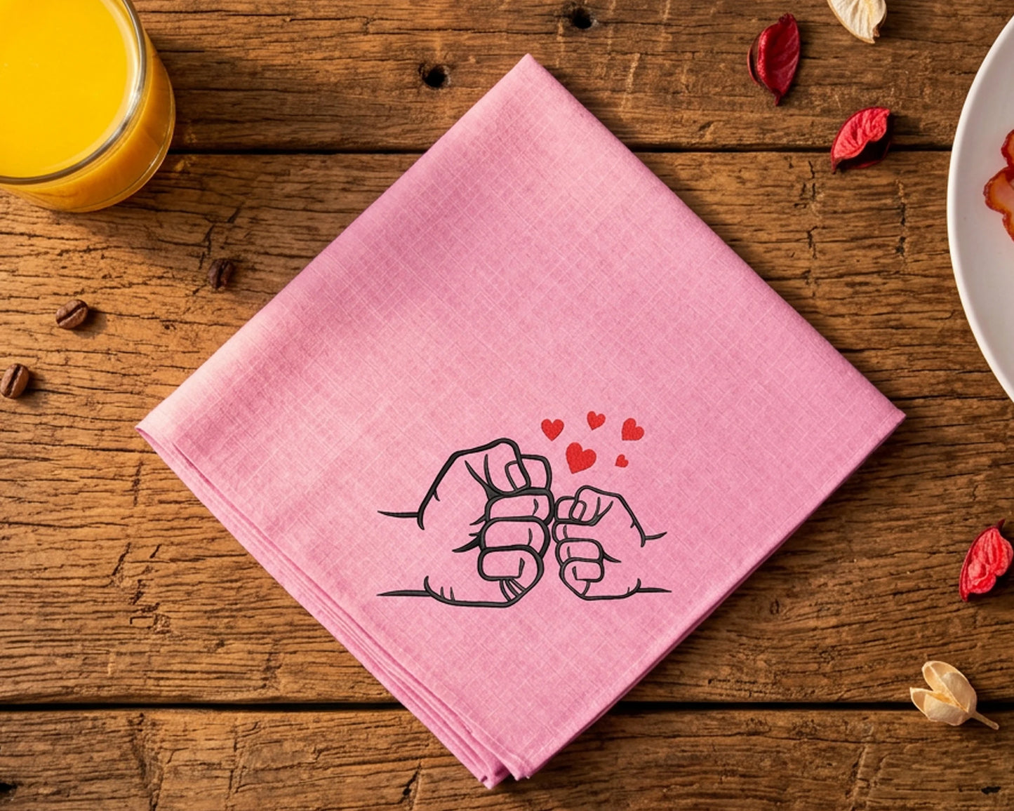 Pink hanky with fists and hearts design on a wooden table with a glass of orange juice and a plate.