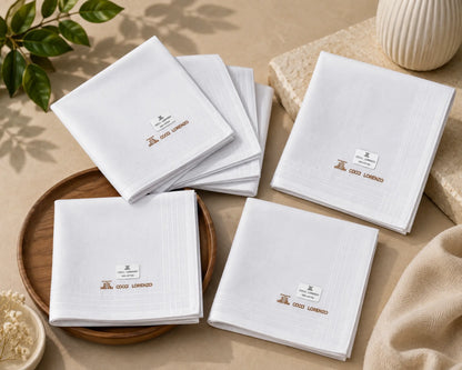 Five folded white linen napkins with gold embroidered logos and small white labels, arranged on a beige tabletop with a wooden tray, stone decor, and a leafy sprig.